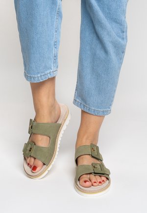 Grüne Wildleder-Sandalen mit zwei breiten Riemen und goldenen Schnallenakzenten. Dicke weiße Sohle, offener Fersenbereich, getragen zu hellblauen, gekürzten Jeans.
