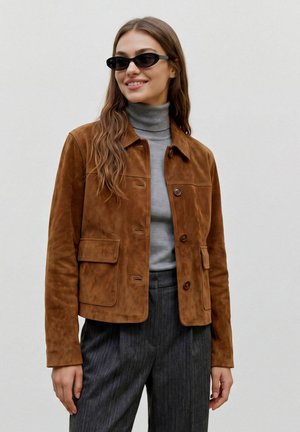 Femme portant des lunettes de soleil noires, une veste en daim marron boutonnée, un pull à col roulé gris et un pantalon gris foncé, souriante sur fond uni.