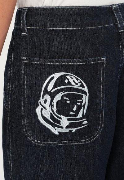 Temno modri kavbojki z belim izvezenim astronavtom na zadnjem žepu. Tkanina ima gladko, trpežno teksturo.