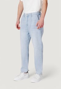 Pantaloni in denim azzurro chiaro con vestibilità rilassata, dotati di una vita elasticizzata, chiusura anteriore con bottone e tasche laterali. Abbinati a scarpe da ginnastica bianche.