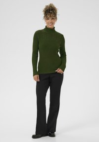 Zaļš adīts turtlenecks džemperis ar teksturētu rakstu ap kaklu, apvienots ar melnām platām biksēm un melnām kurpēm.