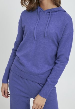 Kobieta ubrana w miękki, fioletowy dzianinowy hoodie i pasujące spodnie, ręce swobodnie wzdłuż ciała, na jednolitym jasnym tle.