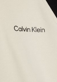 Tessuto color crema con una finitura texturizzata, caratterizzato da un logo Calvin Klein nero ricamato sul lato sinistro superiore.