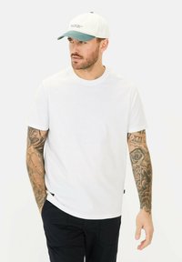 Witte t-shirt met een ronde halslijn, korte mouwen en een ontspannen pasvorm. G gedragen met marine-blauwe broek; het model draagt ook een pet met groene accenten.
