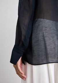 Camisa marinha transparente com mangas longas, punhos abotoados e um design ligeiramente ajustado. O tecido tem uma textura fina com leve translucidez.