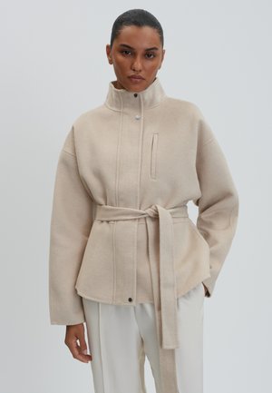 Donna che indossa una giacca di lana beige con cintura, collo alto e pantaloni bianchi, in piedi davanti a uno sfondo chiaro e uniforme.