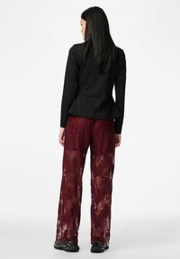 Sieviete ar gariem tumšiem matiem, valkājot melnu garām piedurknēm topu un caurspīdīgas bordo krāsas mežģīņu plašu kāju bikses, stāvot ar muguru pret baltu fonu.