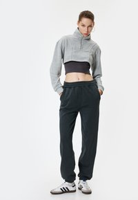 Grijze geknipte sweatshirt met hoge kraag, zwarte strakke laag eronder, donkergroene joggingbroek en witte sneakers met zwarte strepen.