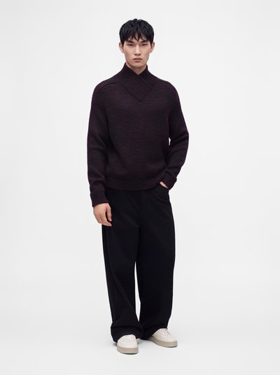 Jeune homme debout portant un pull en maille bordeaux foncé, un pantalon noir ample et des baskets blanches, une main dans la poche.