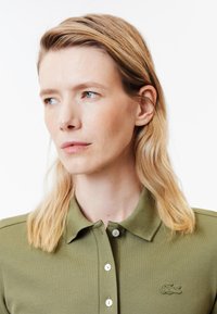 Olivgrön polo med texturerad tyg, med en knappslå och en liten broderad logga på vänster sida. Mjuk krage och korta ärmar.