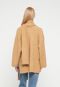 Soe villane kampsun sooja beežis toonis, millel on tekstuuriga palmikmustrit, sobiva salliga, mis on üle õlgade laotatud.