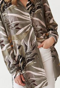 Camicia a maniche lunghe con bottoni decorata con motivi astratti di foglie nei toni del verde, marrone e beige, indossata con pantaloni bianchi, mano in tasca.