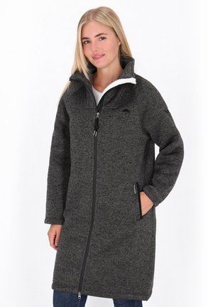 Frau in einem langen, dunkelgrauen Fleece-Mantel mit Reißverschluss, hohem Kragen und Seitentaschen, die steht und gegen einen weißen Hintergrund lächelt.