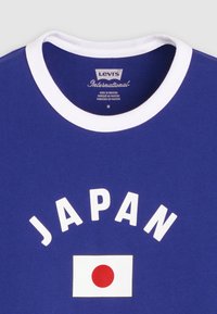 Μπλε μπλούζα με λευκή γιακά, τη λέξη «JAPAN» με λευκά γράμματα και ένα λευκό ορθογώνιο με έναν κόκκινο κύκλο που αντιπροσωπεύει τη σημαία της Ιαπωνίας.