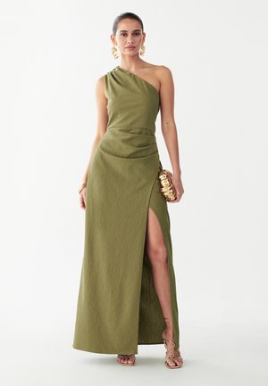 Donna con abito verde oliva monospalla con spacco alto, orecchini dorati, bracciale, pochette e tacchi con cinturini, in piedi su sfondo bianco.
