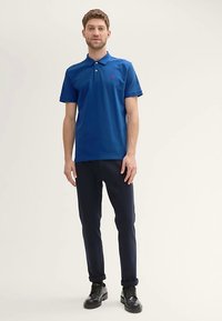 Blå pikétröja med krage och två knappar, kombinerad med marinblå byxor och svarta skor. Enkel design och figurnära silhuett.