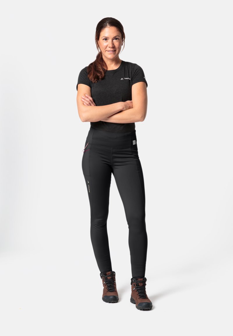 Schwarzes Sportoberteil und eng anliegende schwarze Hose mit Taschen, kombiniert mit braunen Wanderschuhen. Der Stoff wirkt glatt mit funktionalen Designelementen.