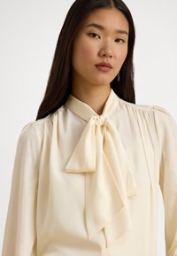 Krém színű blúz masnival a nyaknál, puha anyagból, hosszú ujjak, és finom redőzéssel a vállaknál.