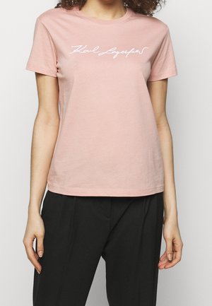 Femme portant un t-shirt à manches courtes rose clair avec l'inscription "Karl Lagerfeld" en blanc sur la poitrine et un pantalon noir, debout devant un fond uni.