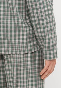 Traje a cuadros verde y blanco con una textura suave. Presenta un corte entallado y mangas largas, mostrando una mano sobre la manga.