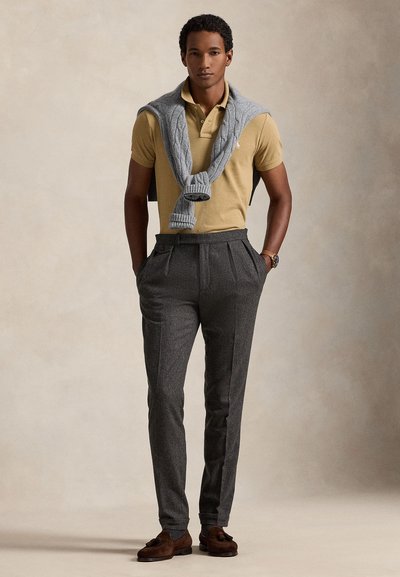 Beige pikétröja, grå stickad tröja över axlarna, grå veckade byxor, bruna mockaskor. Enkel, klassisk design med subtila texturer.