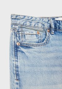 Jean en denim bleu clair en gros plan montrant la taille, la passants de ceinture, la poche avant, la petite poche à monnaie et les rivets en cuivre sur un fond uni.