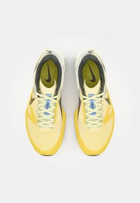 Atletske cipele sa žutim i sivim gornjim dijelom, mrežastom teksturom, i kontrastnim crnim swoosh-om. Otkivači su svijetlosivi s plavim akcentom.