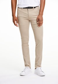 Béžové slim-fit kalhoty s předními kapsami, viditelná textura látky, nošené s černým opaskem a bílými teniskami.