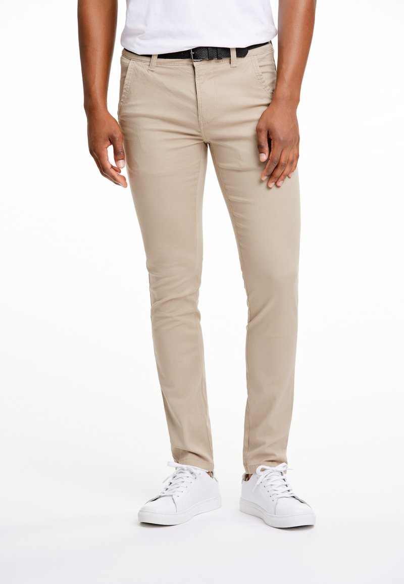 Béžové slim-fit kalhoty s předními kapsami, viditelná textura látky, nošené s černým opaskem a bílými teniskami.