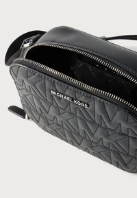 Μαύρο δερμάτινο τσάντα χιαστί με καπιτονέ σχέδιο, ασημένιο φερμουάρ και λογότυπο "Michael Kors" μπροστά, η οποία είναι μερικώς ανοιχτή αποκαλύπτοντας τη εσωτερική επένδυση με το εμπορικό σήμα.