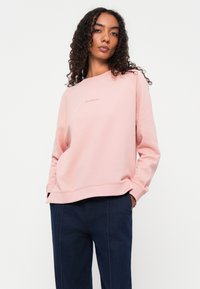 Felpa rosa chiaro con scollatura rotonda e polsini a coste, caratterizzata da un piccolo logo testuale sul davanti. Abbinata a pantaloni blu scuro.