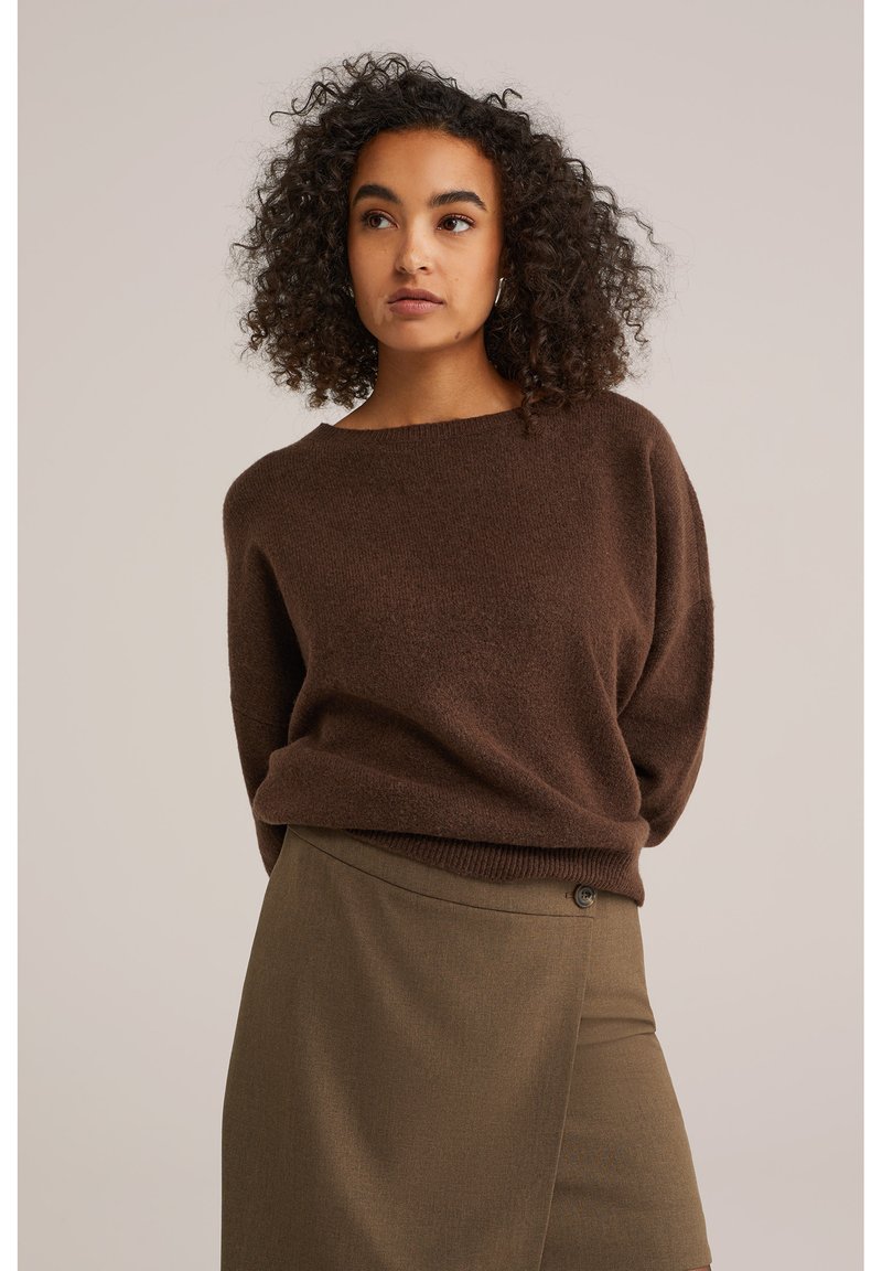 Bruine gebreide trui met een brede halslijn en een relaxte pasvorm, gecombineerd met een bruine rok met een knoopdetail en een subtiele textuur.