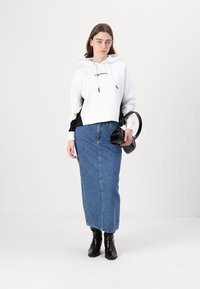 Hanorac scurt alb cu accente negre pe laterale, asociat cu o fustă lungă din denim albastru, botine negre și ținând o geantă de mână neagră.