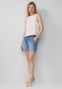 Top sans manches blanc avec des détails en œillets, associé à un short en denim bleu clair et des sandales blanches à lanières, se tenant devant un fond neutre.