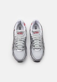 Bílé sportovní boty se stříbrnými akcenty, s prodyšnými panely, kulatou špičkou a šňůrkami. Logo 'ASICS' v červené barvě na jazyku a boku.