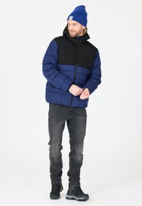Uppblåst jacka med blå kropp och svarta axlar, med huva och dragkedjestängning, i kombination med mörka jeans och svarta vandringskängor.