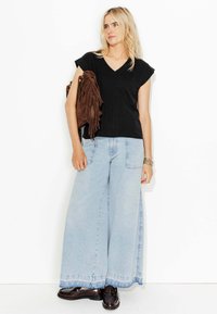 Haut noir à col en V, jeans larges bleu clair avec fentes sur les côtés, sac en cuir frangé marron et mocassins marron foncé. Textures lisses et couleurs contrastées.