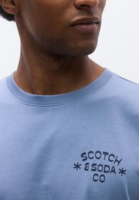 Ανοιχτό μπλε βαμβακερό T-shirt με στρογγυλή λαιμόκοψη και μαύρο λογότυπο κείμενο "SCOTCH & SODA CO" στο κάτω αριστερό μέρος.