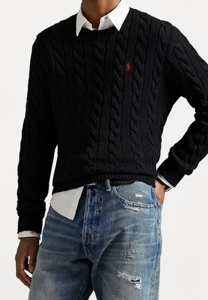 Person wearing black cable-knit sweater with red logo over white button-up shirt, paired with distressed blue jeans, hand in pocket.