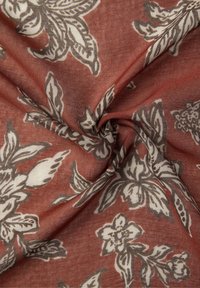 Tessuto di cotone con una base rosso-marrone, caratterizzato da un motivo floreale in tonalità crema e grigio, piegato delicatamente per mettere in evidenza la texture e il design.