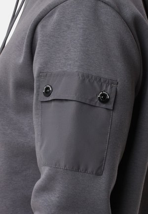 Grijze hoodie met een gladde textuur, met een opgestikte zak op de rechtermouw met drukknoopsluitingen. De stof lijkt zacht en casual.