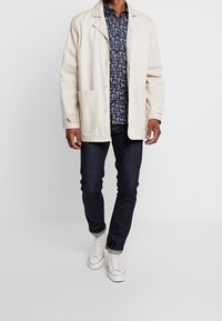 Chaqueta de algodón beige con cierres de botón, dos bolsillos frontales, llevada sobre una camisa de estampado floral azul marino, combinada con vaqueros oscuros y zapatillas blancas.