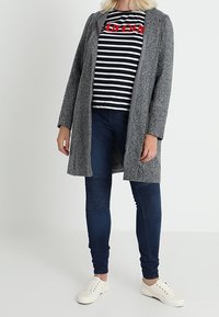 Abrigo gris texturizado sobre una camiseta de rayas en blanco y negro con letras rojas, combinado con jeans azul oscuro y zapatillas blancas.