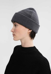 Grijze gebreide beanie met een omgeslagen rand, een gestructureerd oppervlak en een strakke pasvorm, gedragen met een zwarte geribbelde trui. Zijaanzicht.