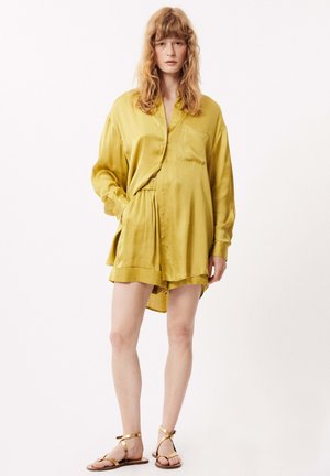 Chemise en satin jaune avec des détails de boutons et une poche, associée à un short assorti. Tissu texturé et design minimaliste. Sandales à lanières marron.