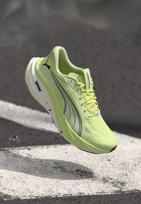 Ανοιχτό πράσινο αθλητικό παπούτσι με χοντρή λευκή σόλα, μαύρο λογότυπο Puma και κίτρινα κορδόνια, εμφανιζόμενο στον αέρα πάνω από επιφάνεια από άσφαλτο.