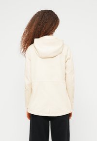 Personne aux cheveux longs et bouclés portant une veste à capuche beige clair et un pantalon noir, vue de dos sur un fond blanc uni.