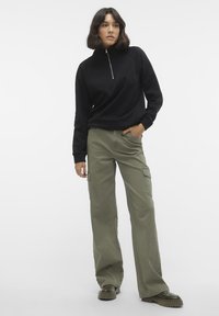 Svart zip-up hettegenser med ribbede mansjetter, paret med olivengrønne cargo-bukser med sidelommer og avslappet passform, iført mørke sko.