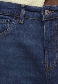 Σκούρο μπλε τζιν τζιν με μπροστινή μέση με κουμπί, θηλιές για ζώνη και κίτρινο ράψιμο, σε συνδυασμό με κίτρινη ριμπ δεμένη μέση πουλόβερ.