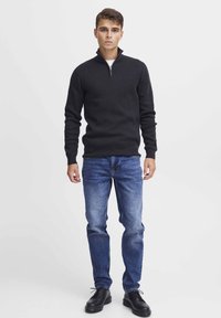 Schwarzer Pullover mit Reißverschluss am Hals und gerippten Bündchen, kombiniert mit hellblauen Jeans und schwarzen Schnürschuhen. Schlichte, körperbetonte Silhouette.